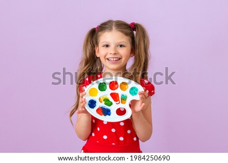 A little girl in a red dress holds a palette with different colors and smiles.