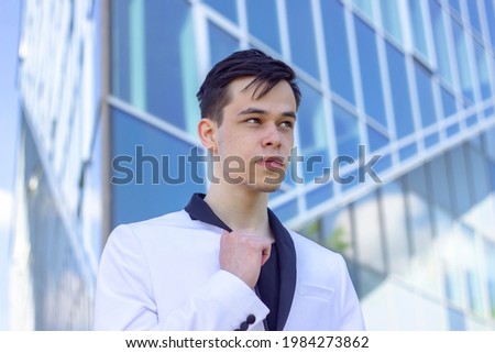 Portrait of a young guy 17-21 years old in a white jacket on the background of glass windows of an office building. Concept: start of training and practice, get a job after college or institute.