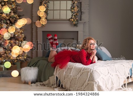 Pretty woman in a Christmas sweater, red skirt and Christmas socks. In the New Year's interior with a Christmas tree decorated with New Year's balls and bulbs. Waiting for the holiday.