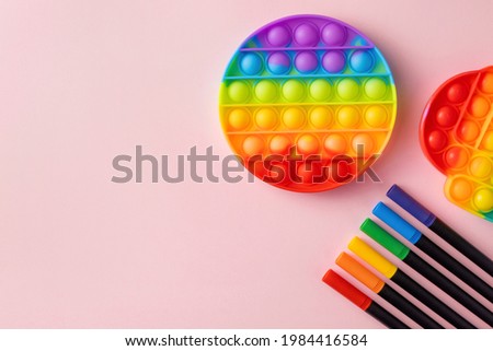 Poppit or Pop it sensory toy and colorful felt pens on pastel pink background. Silicone rainbow bubbles for stress and anxiety relief. Royalty-Free Stock Photo #1984416584