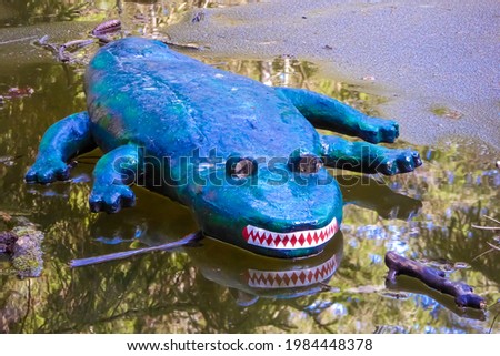 Alligator dummy that swims in a greenish forest lake . Crocodile plastique with white teeth and red tongue . Outdoor art project .