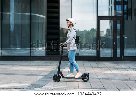 Side view of a young businesswoman riding electric push scooter. Female in cycling helmet driving electrical scooter against building. Royalty-Free Stock Photo #1984449422