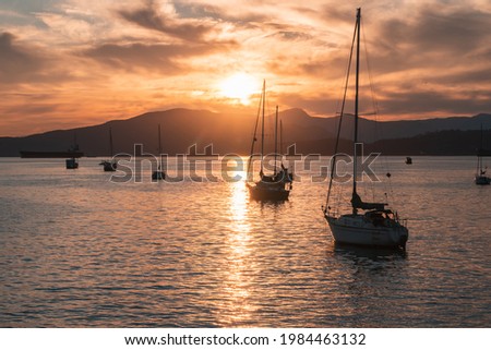 Sailboats sit on calm waters as the sun sets behind a mountain.
