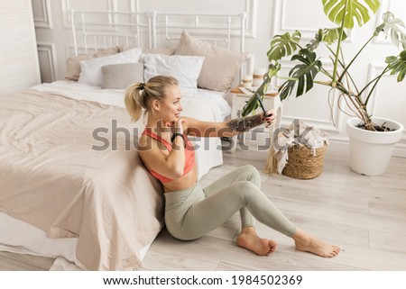 Beautiful female yoga instructor using smartphone, making online workout from home, smiling. Happy young woman in stylish sportswear making a selfie or recording training video, distant work concept