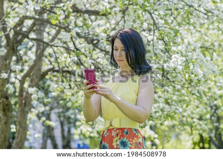 A young woman in a yellow dress stands among a blossoming apple orchard and takes pictures of herself on a mobile phone