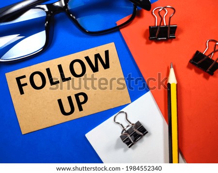 Business concept. Brown card with text FOLLOW UP with pencil,paper clips and glasses on blue,red and white background