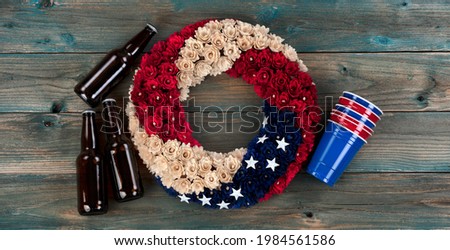 Wreath with US national colors of red, white and blue with stars. Plus, plastic drinking cups and beer on faded blue wooden planks for happy memorial or Independence Day background concept 