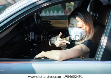 Asian woman in face mask sitting in car with open window, smiling making a shooting gesture to her shoulder covered with bandage plaster after getting Covid-19 vaccine injection via drive-thru service