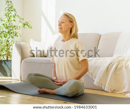Peaceful senior woman in lotus position meditation with closed eyes at home while sitting on yoga mat on floor, full length. Calm elderly lady practicing meditation techniques and yoga indoors Royalty-Free Stock Photo #1984633001