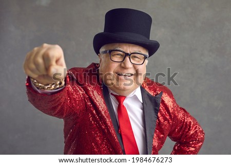 Funny rich mature man in eyeglasses, bling fancy sequin jacket and black top hat smiling and looking at camera. Happy elderly toastmaster, magician, conjurer or millionaire posing on gray background