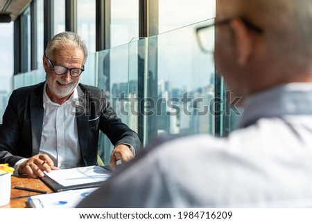 Picture of two business man discussing business and seem like an agreement is made. The young man is wearing white shirt with black tie. The older man is wearing black suit. They meet in hotel lobby.