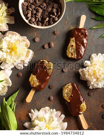 chocolate popsicle with edible gold on a stick on a dark background. Round pieces of chocolate around and in the bowl. Peonies and leaves. Top view. Chocolate ice cream made from dark, milk chocolate