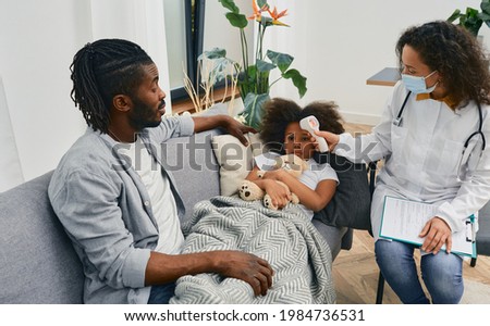House call doctors. Pediatrician measures temperature of a little girl patient with cold and flu using infrared thermometer at home. Medical help at home Royalty-Free Stock Photo #1984736531