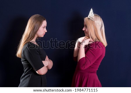 Two young girls, a blonde and a brunette, stand opposite each other. Staring eye to eye. Confrontation. One is wearing a royal diadem.
