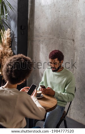 African american man using smartphone near cup of coffee and girlfriend in cafe