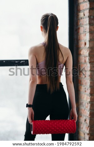 Photo from behind. A lean physique girl in sportswear holds a fascia in her hands while standing against a background of windows