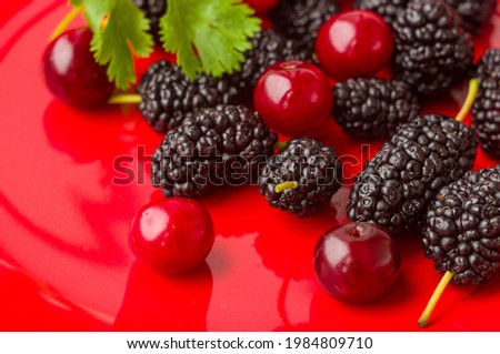 Ripe large black mulberries and red cherries on a red plate, soft focus, close up.