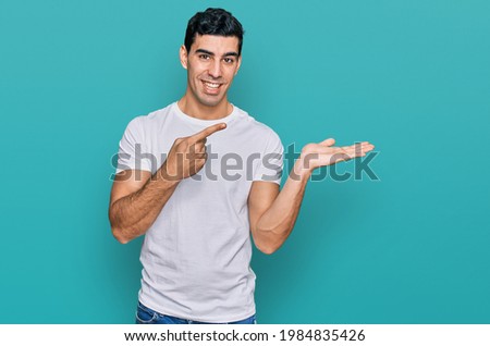 Handsome hispanic man wearing casual white t shirt amazed and smiling to the camera while presenting with hand and pointing with finger. 