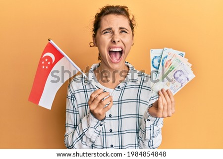Young brunette woman holding singapore flag and dollars angry and mad screaming frustrated and furious, shouting with anger looking up. 