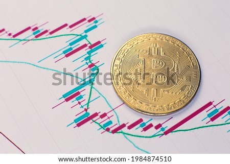 Gold coin of bitcoin cryptocurrency and charts of changes in cryptocurrency rates lie on a wooden table in the office