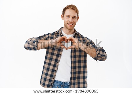 Love you. Caring husband with red hair, showing heart gesture and smiling, express his affection, like someone, standing over white background. Relationship concept