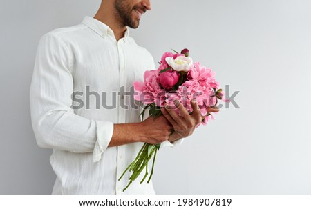 Cropped image of a handsome man holding the bouquet of pink peonies for a gift for valentines or birthday celebration. Smiling male in white shirt holding flowers isolated on the grey background.