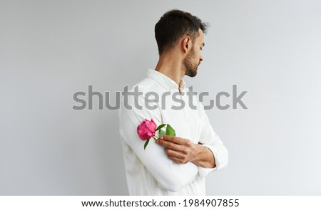 Studio image of a handsome bearded man holding a pink peony in crossed arms. Male wearing white shirt posing with a flower, isolated over grey background.