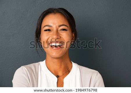 Close up face of beautiful mature woman looking at camera with big laugh. Mid adult hispanic woman standing isolated against grey wall while having fun. Portrait of smiling indian lady laughing. Royalty-Free Stock Photo #1984976798