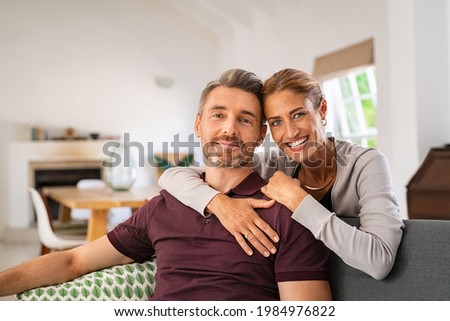 Middle aged couple embracing on sofa while looking at camera. Mature happy woman embracing her husband from behind while relaxing at home. Portrait of smiling wife and man loving in perfect harmony. Royalty-Free Stock Photo #1984976822