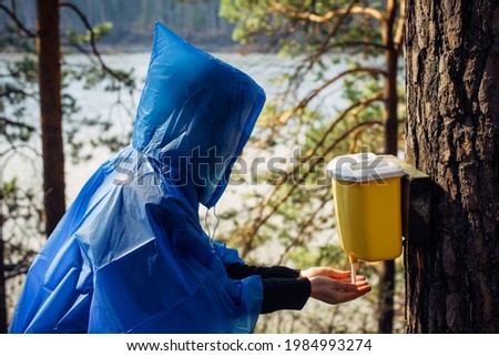 Woman in blue raincoat washes her hands in wash basin hanging on tree. Morning after rain at tourist camp in the forest by the river. Girl's face hidden by hood. Hiking lifestyle.