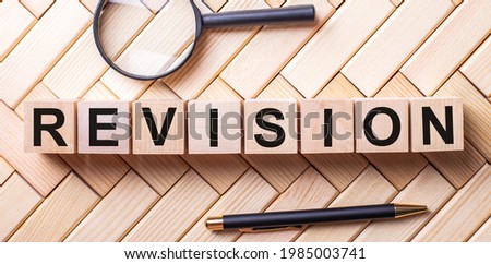 Wooden cubes with the word REVISION stand on a wooden background between a magnifying glass and a pen
