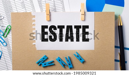 On the desktop are reports, blue clothespins and charts, a pen, a notebook and a sheet of paper with the text ESTATE. Business concept