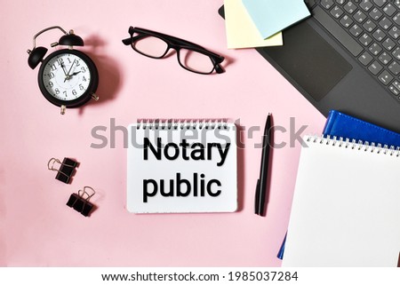 NOTARY PUBLIC is written in a notepad near a laptop, alarm clock, paper and a pen on a pink background. Business concept.