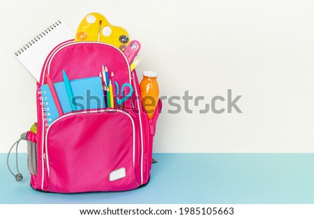 Pink backpack with school supplies standing on white and blue background. Copy space.