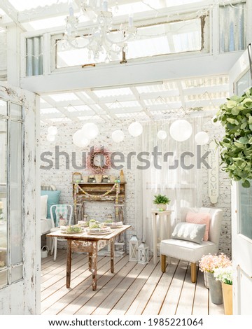 Vintage shabby chic garden room. A white distressed brick wall and antique farm table with diy faux fireplace, chandelier, paper lanterns and linen chair create a playful whimsical space. Royalty-Free Stock Photo #1985221064