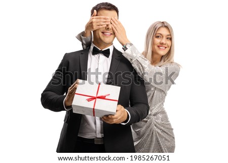 Young woman in elegant dress surprising a man with a present and covering his eyes isolated on white background