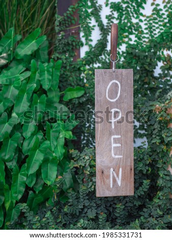 The image of the open signboard on green leaves background focuses on a signboard. The open signboard hanging on an iron pole with green plant leaves in the background. Signboard for small businesses