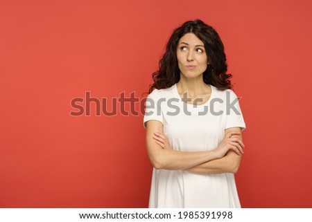 Sarcastic doubtful young woman look with judgmental glance and skeptical face expression side to empty copy space over red wall background. Studio portrait of unsure suspicious female in white t-shirt Royalty-Free Stock Photo #1985391998