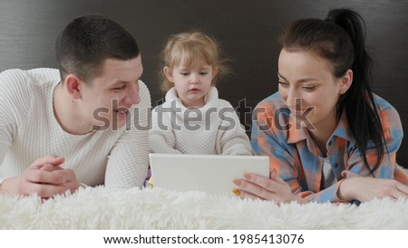 Modern gadget, parents and kid are watching cartoon. Happy family, parents and small child, daughter use tablet together, lying on bed. Mother, child and father are resting at weekend together at home