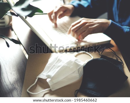Work from home during the COVID-19 pandemic theme double mask over paper file folder with blurred background of male hands using laptop on wooden table with green plant and coffee mug. selective focus