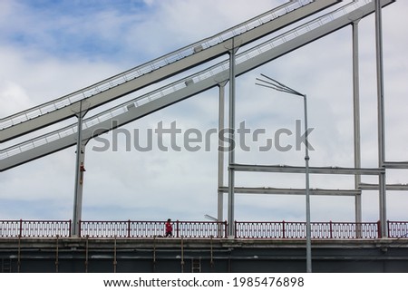 A modern pedestrian iron cable-stayed bridge silhouette with supporting structure, a lantern against a blue cloudy sky in the daytime. A person in a red jacket walking on a bridge. Urban architecture.