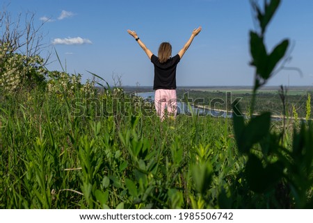 Young woman stands on green hill with view on fields and river, happy woman look in the distance with raised hands to the blue and clear sky. Concept of tourism, freedom, enjoying life in nature.