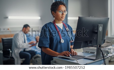 Hospital Ward: Professional Smiling Black Female Head Nurse or Doctor Wearing Stethoscope Uses Medical Computer. In the Background Patients in Beds Recovering Successfully after Sickness and Surgery Royalty-Free Stock Photo #1985507252