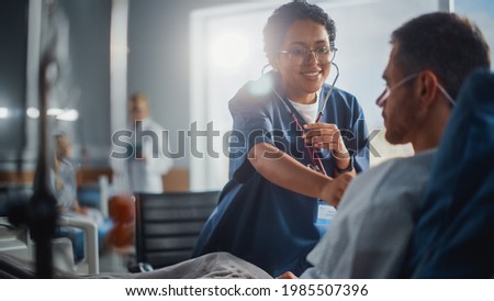 Hospital Ward: Friendly Black Head Nurse Uses Stethoscope to Listen to Heartbeat and Lungs of Recovering Male Patient Resting in Bed, Does Checkup. Man Getting well after Successful Surgery Royalty-Free Stock Photo #1985507396
