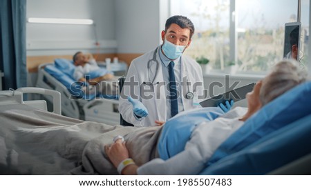 Hospital Ward: Friendly Doctor Wearing Face Mask Talks to Beautiful Senior Female Patient Resting in Bed, Explains Test Results, Gives Recovery Advice. Physician and Old Lady Recovering after Covid-19 Royalty-Free Stock Photo #1985507483