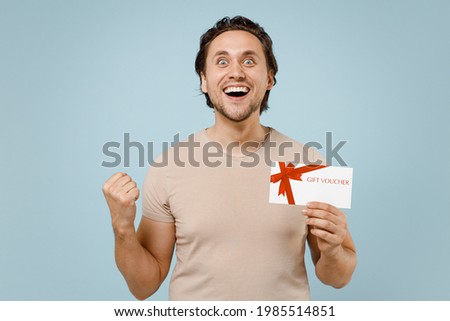 Young excited happy overjoyed caucasian man 20s in casual basic beige t-shirt hold in hands gift voucher flyer mock up clench fist do winner gesture isolated on pastel blue background studio portrait