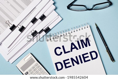 On a light blue background there are documents, glasses, a calculator, a pen and a notebook with the text CLAIM DENIED. Close-up of the workplace. Business concept