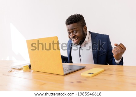 Emotional freelancer sitting at desk at home or office with laptop, smart phone.Young African American entrepreneur dream come true, very excited, wears smart casual suit, celebrates at the office
