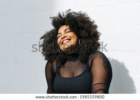 Happy woman in fashionable wear standing near wall. Pretty female in black outfit on white background. Royalty-Free Stock Photo #1985559800