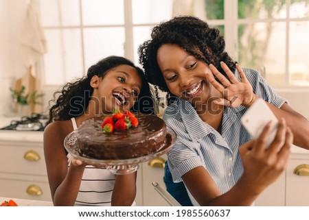 Happy mother and daughter making a video call, with girl holding freshly prepared cake. Woman with her girl holding cake making a video call on her mobile phone.